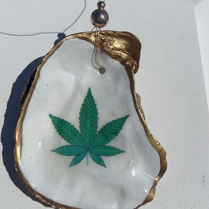 May include: A white oyster shell with a gold rim, featuring a green marijuana leaf design. The shell is strung with a thin wire and a silver bead.