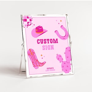 May include: A pink and silver framed sign with a pink background and illustrations of a cowboy hat, a horseshoe, a cactus, a boot, and a disco ball. The sign says "Custom Sign" and "Sophie's Last Rodeo".