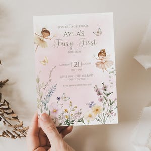 Pu&ograve; includere: Un biglietto d'invito di compleanno rettangolare con illustrazioni ad acquerello di due fate, farfalle e fiori di campo su uno sfondo rosa tenue. Il testo recita: JOIN US TO CELEBRATE AYLA'S Fairy First BIRTHDAY SATURDAY 21 AUGUST 3PM-5PM.
