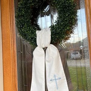 May include: A white fabric wreath bow with a blue cross design. The bow is tied around a green artificial wreath.