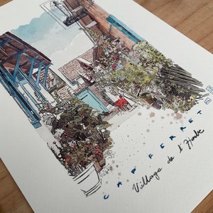 May include: Watercolor painting of a European village scene. The artwork depicts buildings with blue shutters and a red awning, with lush greenery and the text "CAP FERRET Village de l'Herbe".