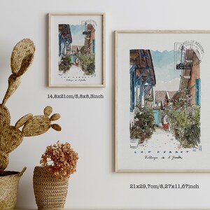 May include: Two framed watercolor prints of a European village scene, one measuring 5.8x8.3 inches and the other 8.27x11.67 inches. The artwork depicts a narrow street with blue accents, lush greenery, and the text "CAP FERRET". A woven cactus and baskets are in the foreground.