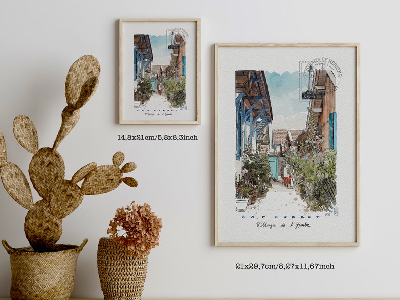 May include: Two framed watercolor prints of a European village scene, one measuring 5.8x8.3 inches and the other 8.27x11.67 inches. The artwork depicts a narrow street with blue accents, lush greenery, and the text "CAP FERRET". A woven cactus and baskets are in the foreground.