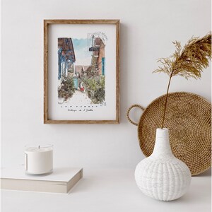 May include: A framed watercolor print of a village street scene with the text "CAP FERRET Village de l'Aiguille". The print is in a wooden frame and is displayed with a white candle, a woven tray, and a white textured vase.