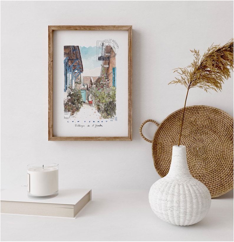 May include: A framed watercolor print of a village street scene with the text "CAP FERRET Village de l'Aiguille". The print is in a wooden frame and is displayed with a white candle, a woven tray, and a white textured vase.
