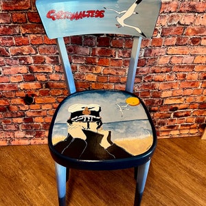 Könnte beinhalten: Ein blauer, bemalter Holzstuhl mit einer Möwe und den Worten "Gorto Maltese" auf der Rückenlehne. Die Sitzfläche des Stuhls ist mit einer Szene bemalt, die eine Person in einem Matrosenhut zeigt, die auf das Meer blickt.