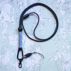 May include: A black and white striped lanyard with a black clip and a black carabiner hook. The lanyard is on a light blue textured background.