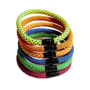 Könnte beinhalten: Ein Stapel aus fünf bunten Seil-Armbändern mit einem schwarzen Magnetverschluss. Die Armbänder sind aus geflochtenem Seil in den Farben Gelb, Orange, Rot, Grün und Blau.