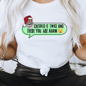 May include: White t-shirt with a green speech bubble that says "Checked it twice and there you are again" with a cartoon Santa Claus and a yellow emoji with a frown.