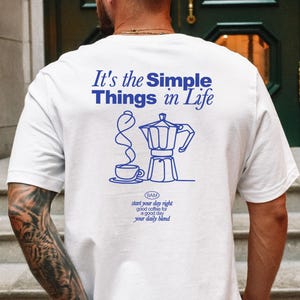 Könnte beinhalten: Weißes T-Shirt mit einer blauen Grafik einer Kaffeekanne und einer Tasse Kaffee. Der Text "It's the Simple Things in Life" befindet sich über der Grafik. Der Text "SAM start your day right good coffee for a good day your daily blend" befindet sich unter der Grafik.