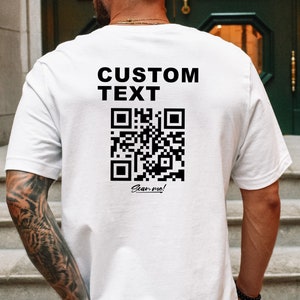 Può includere: T-shirt bianca con la scritta "CUSTOM TEXT" sopra un codice QR nero e le parole "Scan me!" sotto. La maglietta ha maniche corte e un davanti liscio.