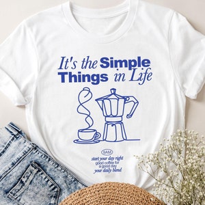 Könnte beinhalten: Weißes T-Shirt mit dem blauen Text "It's the Simple Things in Life". Unter dem Text befindet sich eine blaue Strichzeichnung einer Kaffeemaschine und einer Tasse. Das Shirt wird mit einer Jeans und einem Strohhut kombiniert.