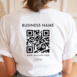 Könnte beinhalten: Weißes T-Shirt mit einem schwarzen QR-Code und dem Text "BUSINESS NAME" darüber. Unter dem QR-Code befindet sich die Website-Adresse "www.yoursite.com" und der Schriftzug "Visit us!". Das T-Shirt hat einen lässigen Stil.