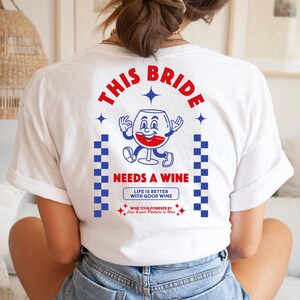 Pode incluir: Camiseta branca com um desenho vermelho e azul. O desenho diz "THIS BRIDE NEEDS A WINE" com um personagem de taça de vinho de desenho animado. A camisa também tem o texto "LIFE IS BETTER WITH GOOD WINE".