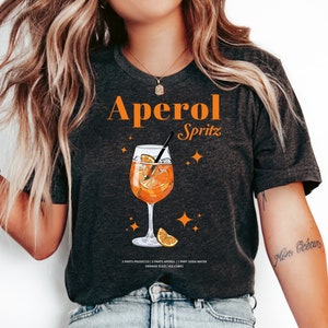 May include: A dark gray t-shirt with the text "Aperol Spritz" in orange letters. The shirt features a graphic of a glass of Aperol Spritz with orange slices and a straw. The text "3 parts prosecco | 2 parts Aperol | 1 part soda water | orange slice | ice cubes" is printed below the graphic.