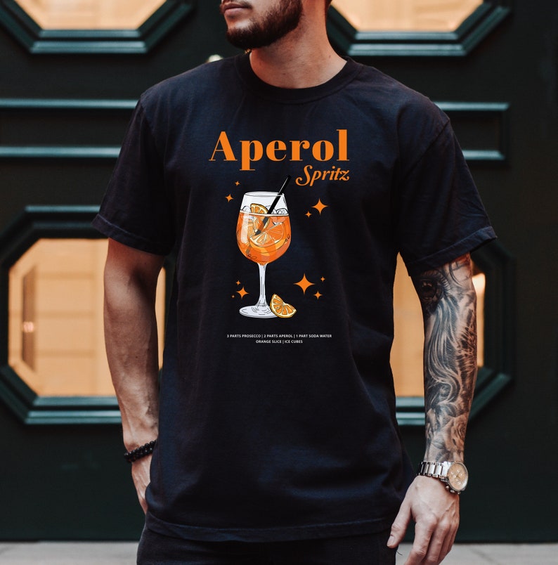 May include: Black t-shirt with a graphic of an Aperol Spritz cocktail. The text "Aperol Spritz" is printed in orange above the image. The recipe for the cocktail is printed below the image.