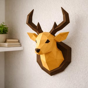 Op de afbeelding: Een geometrische papieren hertshoofd wanddecoratie in tinten geel en bruin. Het hertshoofd is gemonteerd op een donkerbruine zeshoekige plaat. De geweien zijn een donkerdere bruintint. Het kunstwerk wordt tegen een witte muur getoond.