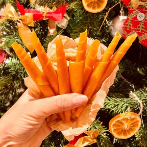 Op de afbeelding: Een bosje oranje fruitleerrolletjes, ook wel fruitrolletjes genoemd, worden in een hand gehouden tegen een achtergrond van een gedecoreerde kerstboom met ornamenten en gedroogde citrusschijfjes.