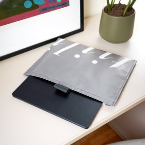 May include: A grey fabric laptop sleeve featuring white stylized text on the front flap. A black rectangular tablet or laptop is partially inserted into the sleeve, which includes a small grey tab closure centered on the edge of the opening.