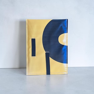 May include: A rectangular yellow pouch featuring a large, dark blue circular graphic design. The pouch includes two vertical dark blue fabric straps attached to the front surface, with visible white stitching along the edges of the material.