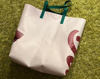 Può includere: Una grande borsa tote color bianco sporco con due manici in tessuto verde scuro. La borsa presenta un design grafico parziale rosso scuro sul lato destro e una piccola parte di un elemento grafico blu e nero visibile sul lato sinistro.