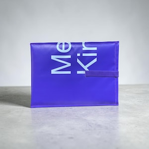 Könnte beinhalten: Eine rechteckige violette Tasche mit dem hellblauen Text Me Kin, der vertikal auf der Vorderseite aufgedruckt ist. Die Tasche verfügt über einen passenden violetten Gummibandverschluss an der rechten Seite, um den Gegenstand sicher zu verschließen.