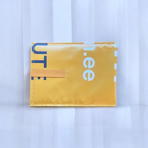 Könnte beinhalten: Eine rechteckige gelbe Tasche mit glatter, synthetischer Textur. Die Vorderseite zeigt teilweise blaue und weiße Schriftzüge mit "UTE" und "n.ee" sowie eine vertikale weiße gestrichelte Linie am rechten Rand. Eine kleine, horizontale gelbe Stofflasche ist angebracht.