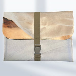 Könnte beinhalten: Eine rechteckige Tasche im Umschlagstil mit einem Druckdesign aus hellbeigen und weißen Stofftexturen. Die Tasche wird durch einen vertikalen olivgrünen Nylongurt mit einer passenden Kunststoff-Steckschnalle auf der Vorderklappe verschlossen.