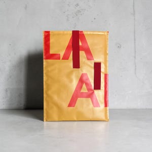 Könnte beinhalten: Eine rechteckige goldfarbene Tasche mit großen, kräftigen roten Buchstaben, die LA AI bilden. Das Design umfasst einen vertikalen roten Stoffriemen, der mittig an der Oberkante angebracht ist, sowie ein kleines rotes rechteckiges Akzentstück.