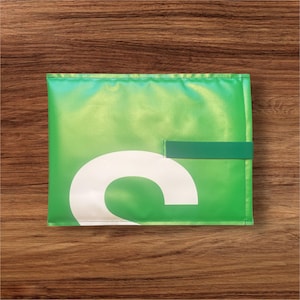 Könnte beinhalten: Eine rechteckige grüne Tasche mit glatter, reflektierender Oberfläche. Auf der Vorderseite befindet sich ein großer, weißer, kleingeschriebener Buchstabe c auf der unteren linken Seite. Ein dunkelgrüner horizontaler Riemen dient als Verschluss.