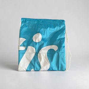 May include: A bright turquoise drawstring backpack made of a smooth, lightweight synthetic fabric. The front of the bag features a large, white graphic print of the letters 'is' in a bold, lowercase, sans-serif typeface with a dot above the letter i.