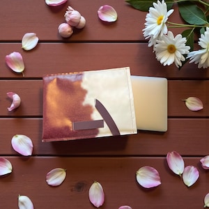Puede incluir: Una cartera beige y marrón con un acento marrón oscuro y un objeto rectangular de color claro. Pétalos de flores rosas y margaritas blancas con centros amarillos están esparcidos sobre una superficie de madera marrón.