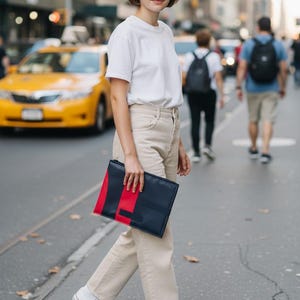 Puede incluir: Un bolso de mano rectangular con exterior azul marino oscuro, una llamativa franja vertical roja y un detalle en negro. El bolso se sostiene en posición horizontal y presenta un diseño limpio y estructurado con una cremallera visible en la parte superior.
