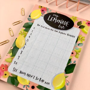 Puede incluir: Un bloc de notas rosa con un diseño floral con limones, flores y hojas. El bloc de notas tiene un círculo negro con el texto "my LEMONADE list" y un área cuadriculada con el texto "TEN GOOD THINGS THAT HAVE HAPPENED RECENTLY" y líneas numeradas del 1 al 10. La parte inferior del bloc de notas tiene el texto "SEE THINGS AREN'T SO BAD XXX".