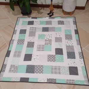 May include: A white and grey patchwork quilt with a mint green border. The quilt is made up of squares of different fabrics, including stars, polka dots, and geometric patterns.