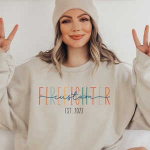 May include: A light beige sweatshirt with the text "FIREFIGHTER custom EST. 2023" in rainbow colours. The text is in a vertical stacked format with the word "custom" written in a cursive font.