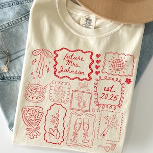 K&ouml;nnte beinhalten: Elfenbeinfarbenes T-Shirt mit rotem Grafikdesign. Das Design enth&auml;lt den Text "future Mrs. Johnson est. 2025" sowie verschiedene Illustrationen wie Kuchen, Ring und Sektgl&auml;ser. Das T-Shirt ist gefaltet.