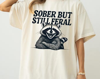 May include: Ivory-colored t-shirt with the text "SOBER BUT STILL FERAL" above a graphic of a smiling raccoon with crossed arms. The raccoon is black and white. The shirt is a casual, oversized fit.