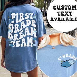 May include: A light blue t-shirt with the text "First Grade Dream Team" in white with a retro font and clouds and stars. The text "Custom Text Available!" is in a black circle.