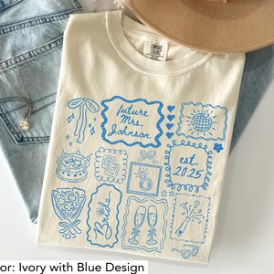 K&ouml;nnte beinhalten: Elfenbeinfarbenes T-Shirt mit blauem Grafikdesign. Das Design enth&auml;lt den Text "future Mrs. Johnson" und "est. 2025" sowie Illustrationen von Kuchen, Sektgl&auml;sern und einem Ring. Das Shirt ist aus weichem Material.