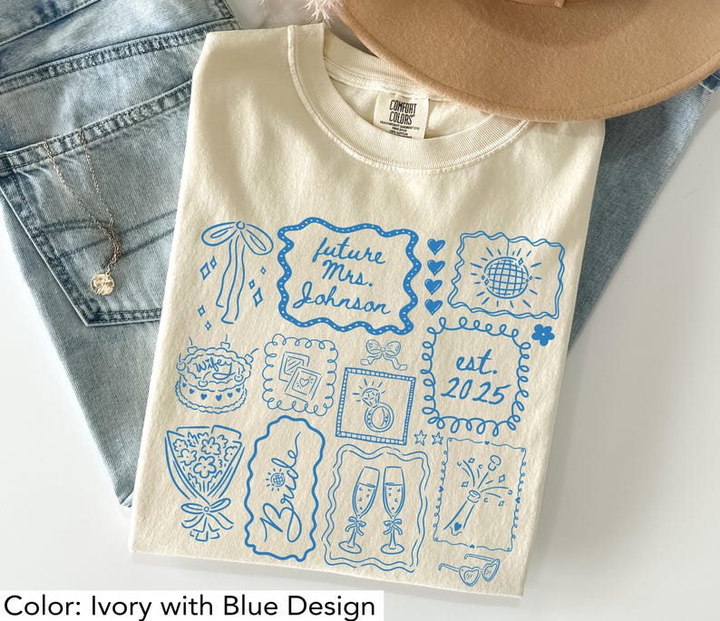 K&ouml;nnte beinhalten: Elfenbeinfarbenes T-Shirt mit blauem Grafikdesign. Das Design enth&auml;lt den Text "future Mrs. Johnson" und "est. 2025" sowie Illustrationen von Kuchen, Sektgl&auml;sern und einem Ring. Das Shirt ist aus weichem Material.