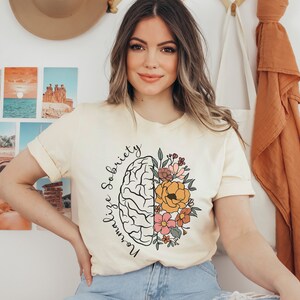 May include: Cream-colored t-shirt featuring a stylized brain graphic with floral elements. The design includes the words "Normalize Sobriety" in a script font. The shirt has rolled-up sleeves and a relaxed fit, suitable for casual wear.