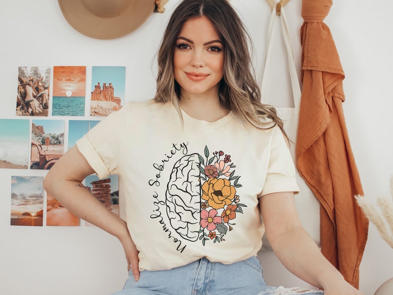 May include: Cream-colored t-shirt featuring a stylized brain graphic with floral elements. The design includes the words "Normalize Sobriety" in a script font. The shirt has rolled-up sleeves and a relaxed fit, suitable for casual wear.