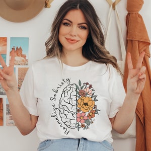 May include: White t-shirt with a graphic design featuring a brain illustration and colorful flowers. The text "Normalize Sobriety" is written in a curved font around the brain. The shirt is a casual style.