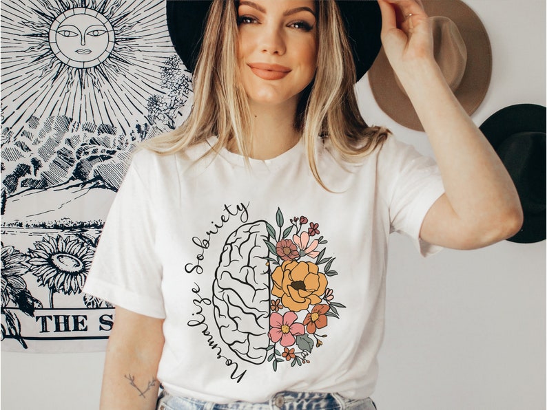 May include: White t-shirt featuring a graphic design of a brain split in half, with one side depicting a brain and the other side filled with colorful flowers. The words "Normalize Sobriety" are written in a curved font around the brain. The shirt is a casual style.