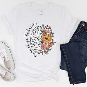 May include: White t-shirt with a graphic of a brain and flowers. The text "Normalize Sobriety" is written around the brain. The shirt is paired with dark blue jeans and white sneakers.