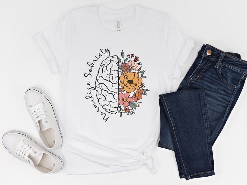 May include: White t-shirt with a graphic of a brain and flowers. The text "Normalize Sobriety" is written around the brain. The shirt is paired with dark blue jeans and white sneakers.