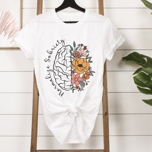 May include: White t-shirt featuring a graphic of a brain split in half, with one side a brain outline and the other side filled with colorful flowers. The words "Normalize Sobriety" are written in a script font around the brain.