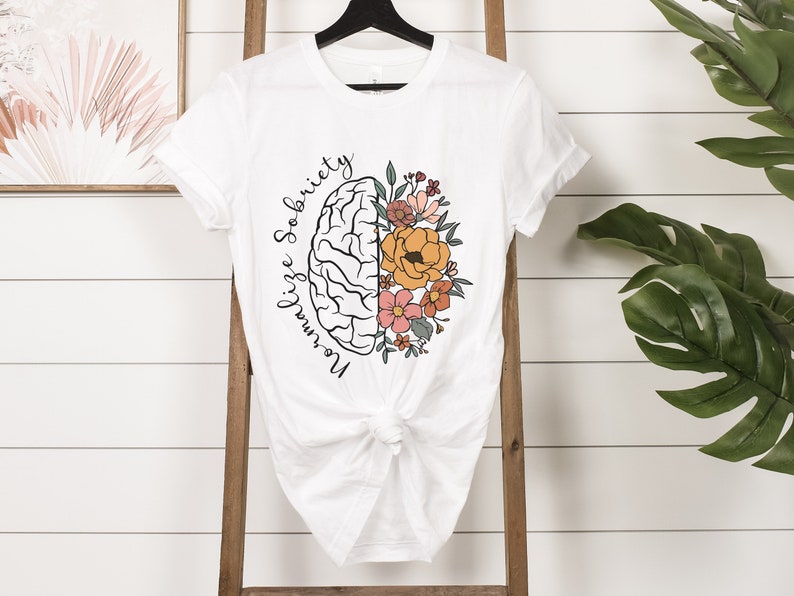 May include: White t-shirt featuring a graphic of a brain split in half, with one side a brain outline and the other side filled with colorful flowers. The words "Normalize Sobriety" are written in a script font around the brain.