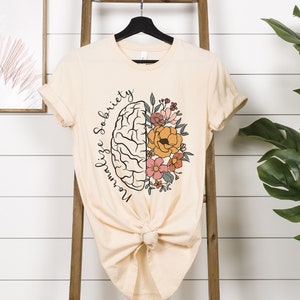 May include: Cream-colored t-shirt featuring a graphic of a brain split in half, with one side a brain and the other side flowers. The text "Normalize Sobriety" is written in a script font around the brain. The shirt is knotted at the bottom.